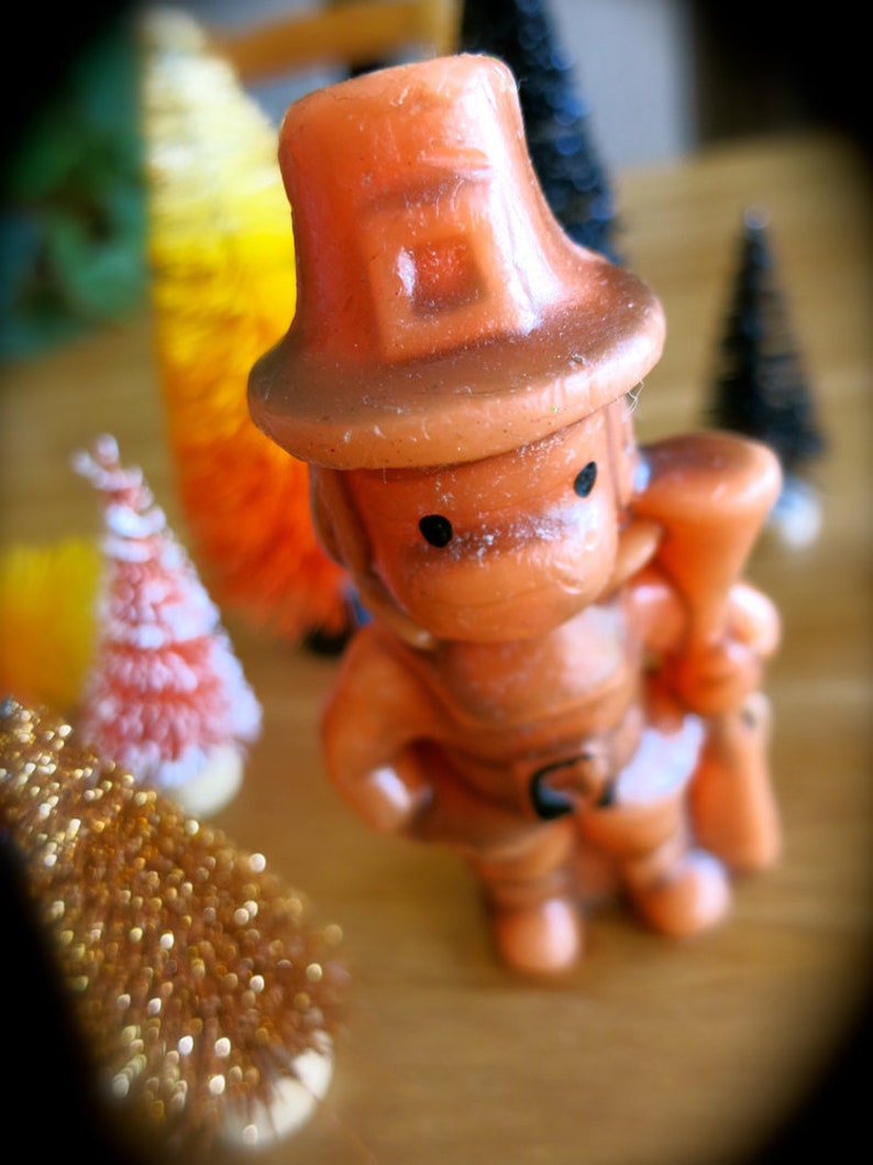 May include: A small, orange figurine of a person wearing a hat and holding a small, brown object. The figurine is made of plastic and has a whimsical, cartoonish style.