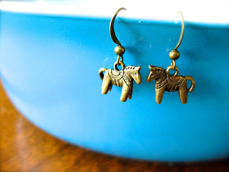 Puede incluir: Un par de pendientes de color bronce con peque&ntilde;os dijes de caballo. Los dijes de caballo tienen dise&ntilde;os de silla de montar detallados y est&aacute;n suspendidos de ganchos para la oreja curvados. Los pendientes est&aacute;n sobre un fondo azul borroso.