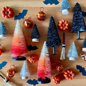 K&ouml;nnte beinhalten: Eine Sammlung von Halloween-Dekorationen, darunter Miniatur-Flaschenb&uuml;rstenb&auml;ume in Schwarz, Wei&szlig; und Orange, kleine K&uuml;rbisse und Kupferglocken mit einem K&uuml;rbisgesicht.