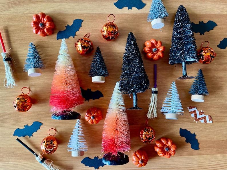 K&ouml;nnte beinhalten: Eine Sammlung von Halloween-Dekorationen, darunter Miniatur-Flaschenb&uuml;rstenb&auml;ume in Schwarz, Wei&szlig; und Orange, kleine K&uuml;rbisse und Kupferglocken mit einem K&uuml;rbisgesicht.