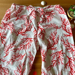 Könnte beinhalten: Weiße Schlafanzughose mit rotem Korallenriffmuster. Die Hose hat einen Kordelzug in der Taille. Das Bild befindet sich auf einer Holzoberfläche mit dekorativen Elementen, darunter eine kleine Sukkulentenpflanze, Muscheln und Ananas-Ausschnitte aus Papier.