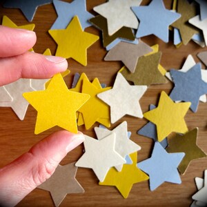 K&ouml;nnte beinhalten: Eine Hand h&auml;lt einen gelben Papierstern &uuml;ber einer Streuung von Papiersternen in verschiedenen Farben, darunter Wei&szlig;, Gold und Blau.