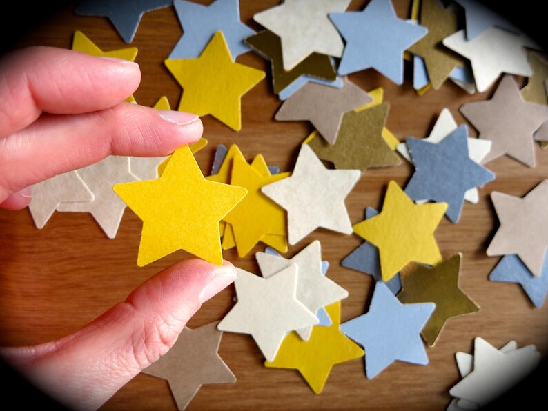 K&ouml;nnte beinhalten: Eine Hand h&auml;lt einen gelben Papierstern &uuml;ber einer Streuung von Papiersternen in verschiedenen Farben, darunter Wei&szlig;, Gold und Blau.