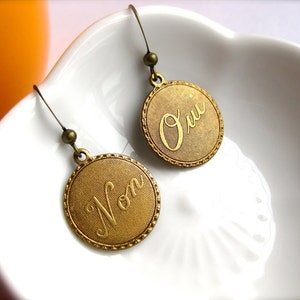 May include: A pair of antique gold-colored earrings. Each earring features a round pendant with a textured surface and a decorative border. One earring says "Non" and the other says "Oui" in elegant script.