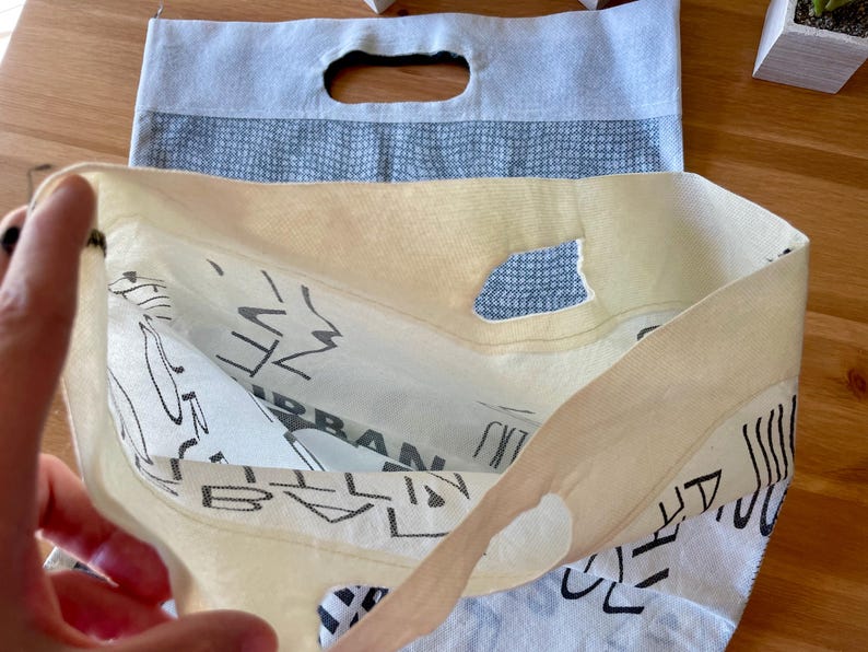 May include: A white canvas tote bag with black text that reads "URBAN" and "CITY" on the front. The bag is folded in half, revealing a blue and white patterned lining.