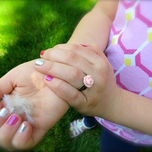 May include: A child's hand wearing a silver ring with a pink flower design. The hand also has pink and silver nail polish.