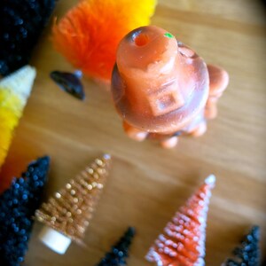 May include: A collection of miniature Christmas trees with various colors and textures, including orange, gold, red, and black. The trees are decorated with glitter and white flocking. There is also a small orange and brown ceramic figurine.