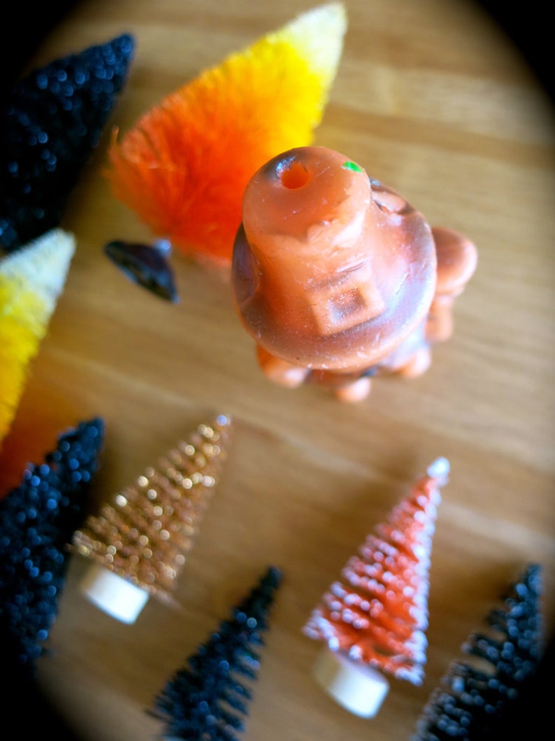 May include: A collection of miniature Christmas trees with various colors and textures, including orange, gold, red, and black. The trees are decorated with glitter and white flocking. There is also a small orange and brown ceramic figurine.