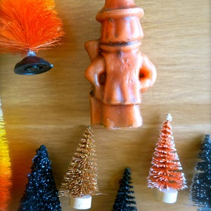 May include: A collection of miniature Christmas trees in various colors, including black, orange, and gold. The trees are decorated with glitter and are arranged on a wooden surface. A small orange figurine of a man wearing a hat is also visible in the image.
