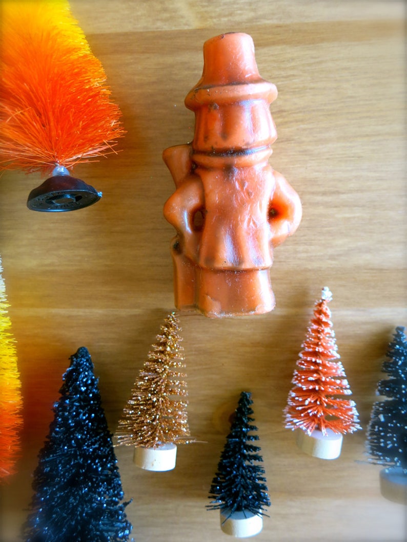 May include: A collection of miniature Christmas trees in various colors, including black, orange, and gold. The trees are decorated with glitter and are arranged on a wooden surface. A small orange figurine of a man wearing a hat is also visible in the image.