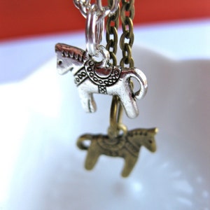 Puede incluir: Primer plano de dos collares con colgantes de caballo. Un colgante es plateado, el otro es de color bronce. Ambos colgantes de caballo est&aacute;n suspendidos de cadenas. El fondo es una mezcla borrosa de rojo y blanco.