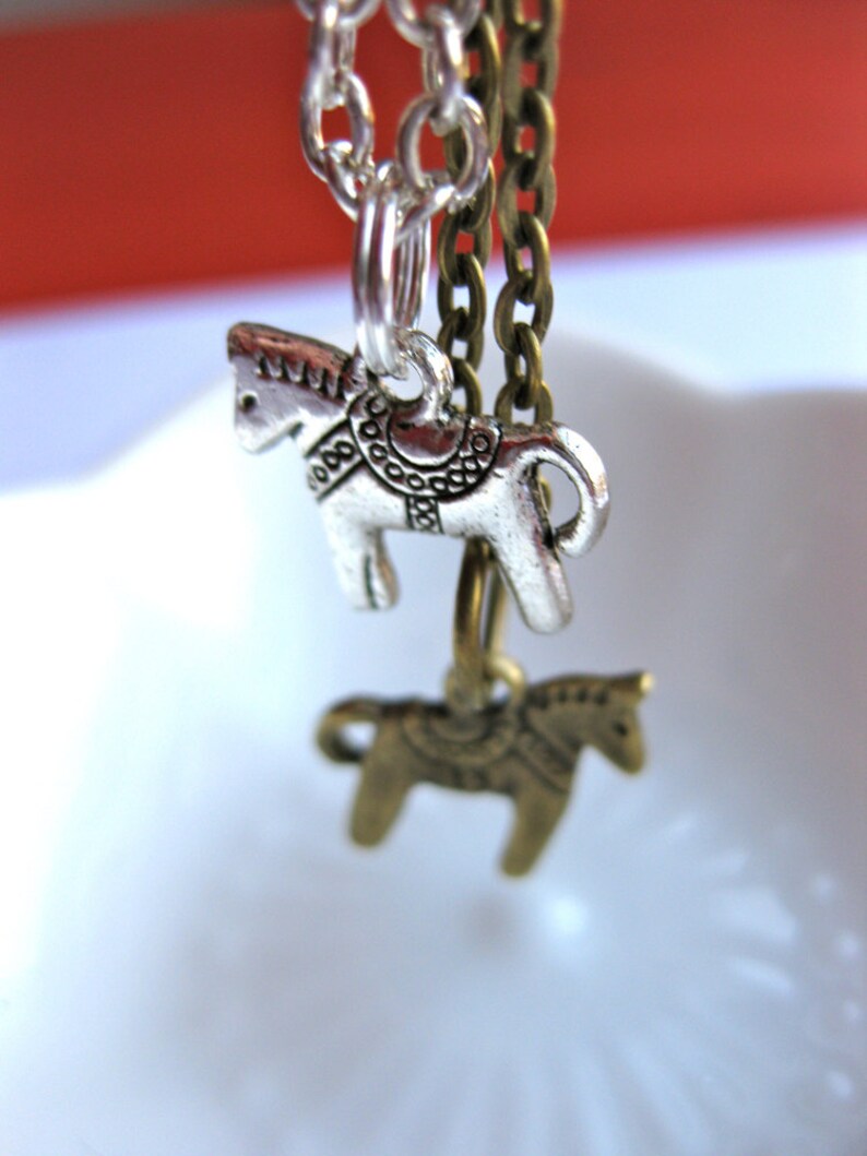 Puede incluir: Primer plano de dos collares con colgantes de caballo. Un colgante es plateado, el otro es de color bronce. Ambos colgantes de caballo est&aacute;n suspendidos de cadenas. El fondo es una mezcla borrosa de rojo y blanco.