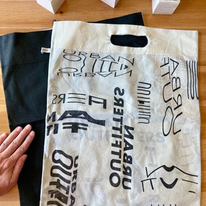 May include: A white canvas tote bag with black text that reads "Urban Outfitters" repeated multiple times. The bag is folded in half and a black tote bag is visible underneath.