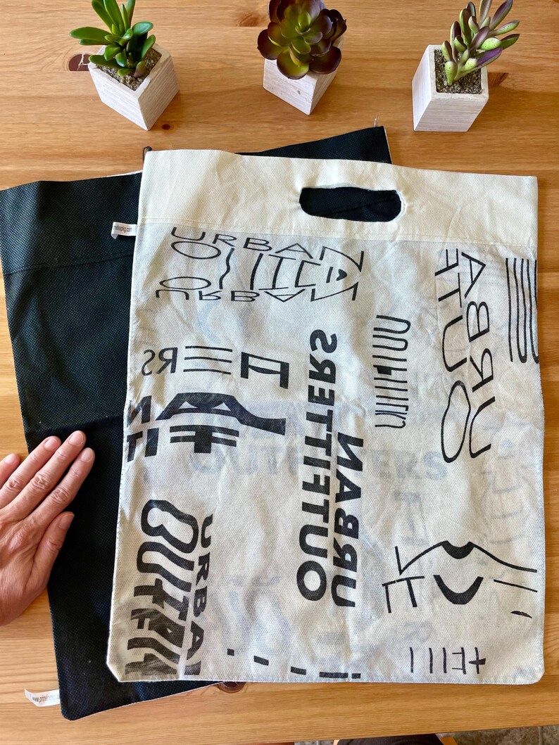May include: A white canvas tote bag with black text that reads "Urban Outfitters" repeated multiple times. The bag is folded in half and a black tote bag is visible underneath.