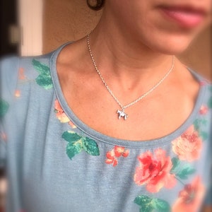 Puede incluir: Un collar de plata con un peque&ntilde;o colgante de caballo. El collar se lleva sobre una camiseta azul claro con estampado floral de rosas rojas y rosas y hojas verdes.