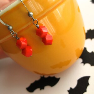 K&ouml;nnte beinhalten: Ein Paar Ohrringe mit orangefarbenen Glasperlen, die an silbernen Haken h&auml;ngen. Die Ohrringe werden auf einer gelben Keramikschale pr&auml;sentiert.