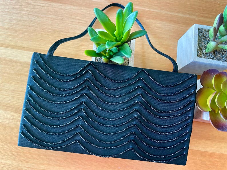 May include: A black satin evening bag with a beaded wave pattern. The bag has a long strap and is closed with a magnetic clasp.