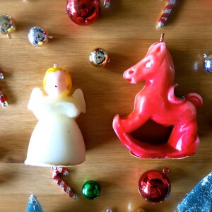 May include: A red rocking horse candle and a white angel candle with gold accents. The candles are surrounded by Christmas ornaments, including red and gold baubles, candy canes, and small green and blue trees.