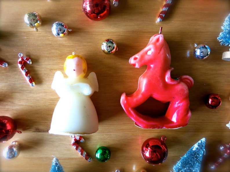 May include: A red rocking horse candle and a white angel candle with gold accents. The candles are surrounded by Christmas ornaments, including red and gold baubles, candy canes, and small green and blue trees.