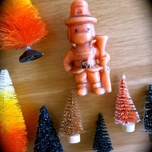 May include: A collection of miniature artificial Christmas trees in various colors, including orange, black, gold, and white. A small orange figurine of a firefighter holding a hose is also included.