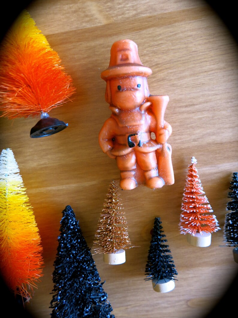 May include: A collection of miniature artificial Christmas trees in various colors, including orange, black, gold, and white. A small orange figurine of a firefighter holding a hose is also included.