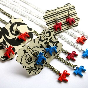 Puede incluir: Una colecci&oacute;n de collares y pendientes con peque&ntilde;os colgantes de caballos de colores. La joyer&iacute;a incluye caballos rojos y azules, con cadenas plateadas y bronce. Los pendientes se exhiben en etiquetas a rayas y estampadas.