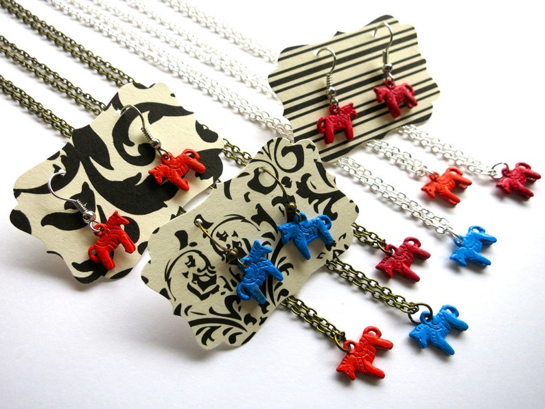 Puede incluir: Una colecci&oacute;n de collares y pendientes con peque&ntilde;os colgantes de caballos de colores. La joyer&iacute;a incluye caballos rojos y azules, con cadenas plateadas y bronce. Los pendientes se exhiben en etiquetas a rayas y estampadas.