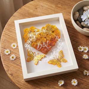 May include: A framed paper art piece featuring a heart made of yellow and orange roses, butterflies, and the letters "B&L". The white-framed artwork sits on a wooden table with daisies and a bowl of stones and flowers.
