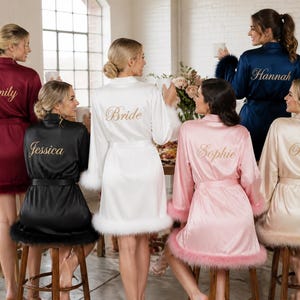 May include: A collection of six satin robes in burgundy, black, white, pink, and navy blue, each featuring long sleeves with feathered cuffs and gold embroidered names or the word Bride on the back, worn by individuals sitting on wooden stools.