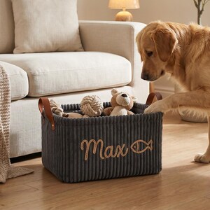 May include: A rectangular dark gray corduroy storage basket featuring brown leather handles and the embroidered name Max next to a fish icon. The basket is filled with various plush toys and rope balls, sitting on a wooden floor.