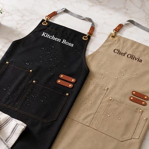 May include: Two aprons, one black and one tan, with the text "Kitchen Boss" and "Chef Olivia" printed on them. Each apron has two front pockets, brown leather accents, and a gray neck strap. The aprons are designed for kitchen use.
