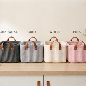 May include: Four square storage baskets with a ribbed texture, each featuring two brown faux leather handles secured with metal rivets. The baskets are arranged in a row and labeled as charcoal, grey, white, and pink, showcasing their distinct colors.