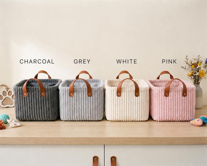 May include: Four square storage baskets with a ribbed texture, each featuring two brown faux leather handles secured with metal rivets. The baskets are arranged in a row and labeled as charcoal, grey, white, and pink, showcasing their distinct colors.