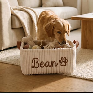 May include: A rectangular off-white corduroy storage basket with brown leather handles. The front features the embroidered name Bean in brown script lettering next to a brown paw print icon. The basket is filled with various dog toys.
