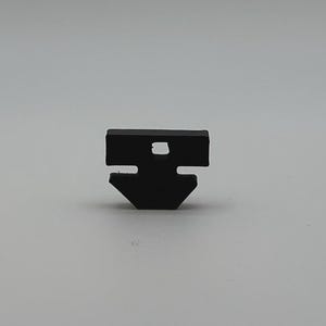 May include: A small, black, plastic component with a unique shape. It features a rectangular top with a square hole and a downward-pointing, angled bottom section. The object is set against a plain, light gray background.