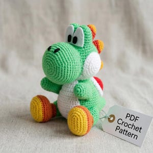 Può includere: Un giocattolo di dinosauro verde all'uncinetto fatto a mano con dettagli bianchi, grandi occhi bianchi e dettagli arancioni e rossi. Il giocattolo ha un'etichetta con la scritta "PDF Crochet Pattern". Il giocattolo è su una superficie neutra.