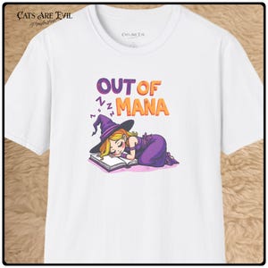 May include: A white short-sleeve t-shirt featuring a graphic of a blonde witch in a purple dress and hat sleeping on an open book. Above the illustration, the text reads OUT OF MANA in purple and orange letters with small purple Zs nearby.