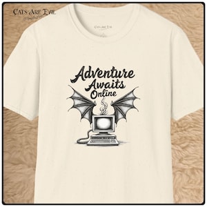 Könnte beinhalten: Ein cremefarbenes Kurzarm-T-Shirt mit gesprenkelter Textur. Auf der Vorderseite befindet sich eine schwarze Grafik eines Vintage-Computers mit Fledermausflügeln und Rauch, der aus dem Monitor aufsteigt. Darüber steht der Text Adventure Awaits Online.
