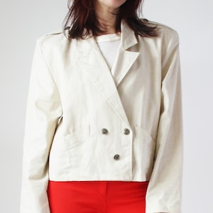 Pode incluir: Um blazer branco de dupla abotoadura com comprimento cropped e dois botões prateados. O blazer é usado sobre uma camiseta branca e combinado com calças vermelhas brilhantes e saltos pretos de bico fino.