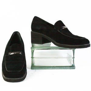 May include: A pair of black suede loafers with a silver-tone buckle detail. One shoe is on a small block, and the other is next to it. The shoes have a low block heel and a square toe.