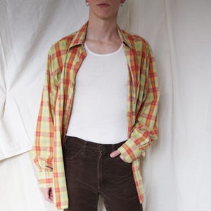 May include: A casual outfit featuring a white undershirt, brown corduroy pants, and a plaid button-down shirt in shades of yellow, orange, and green. The outfit is completed with white sneakers.