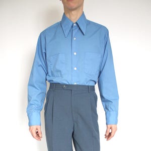 May include: A person wearing a light blue long-sleeved shirt with two chest pockets and a pointed collar. The shirt is paired with grey-blue pleated trousers and black dress shoes. The outfit is suitable for formal or business attire.