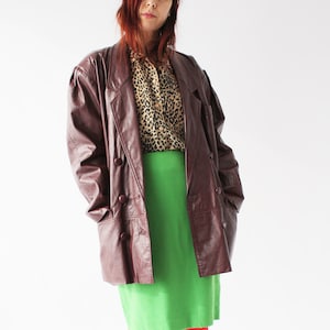 Peut inclure: Une femme portant une veste en cuir marron sur un chemisier à imprimé léopard et une jupe verte. Elle porte des collants rouges et des bottines noires.