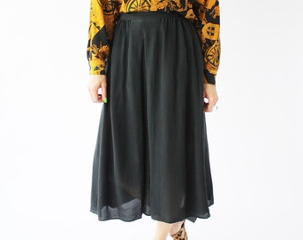 Op de afbeelding: Een zwarte midi-rok met een lang mouwig shirt met een gouden en zwart patroon. Het shirt heeft een kraag en een knoopsluiting aan de voorkant. De persoon draagt ​​schoenen met luipaardprint.