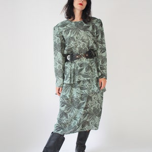 May include: A long, green dress with a black floral print. The dress has a tiered skirt and a black leather belt with a silver buckle. The model is wearing black cowboy boots.