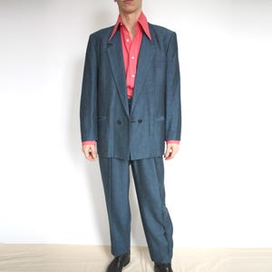 May include: A vintage denim-style suit, with a double-breasted jacket and matching trousers. The suit is worn with a bright pink button-down shirt. The outfit is completed with black leather shoes. The suit is a classic style.