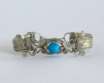 May include: A silver bracelet with an intricate design and a turquoise blue gemstone in the center.