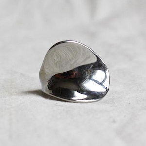 Könnte beinhalten: Ein polierter Silberring mit einer glatten, unregelmäßigen Oberfläche. Der Ring reflektiert das Licht und erzeugt Glanzlichter und Schatten. Das Design ist minimalistisch und modern, geeignet für den Alltag.