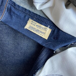 May include: Close-up of a pair of dark blue denim jeans. The interior of the jeans is visible, with a white fabric lining. A rectangular tag displays the "Wrangler" brand name, along with size and care instructions. The jeans are labeled as 28x33.