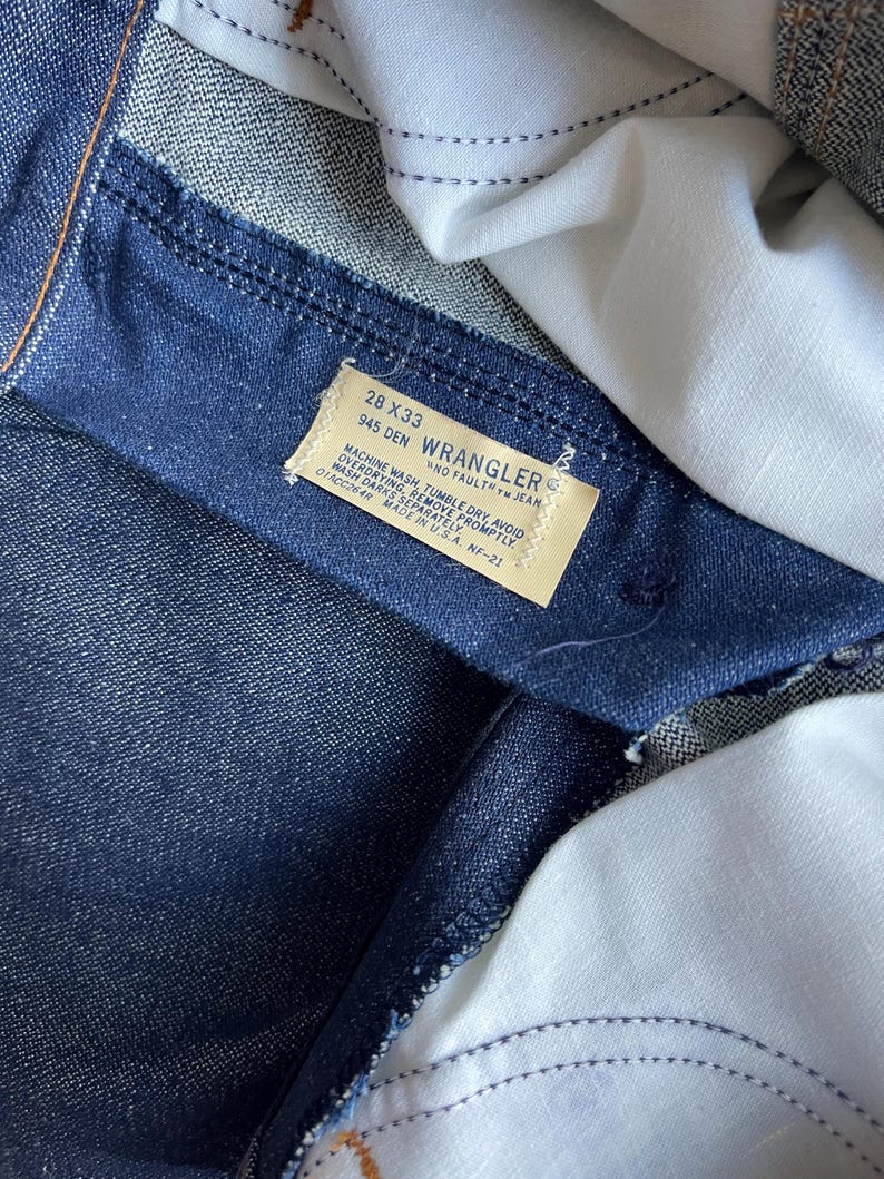 May include: Close-up of a pair of dark blue denim jeans. The interior of the jeans is visible, with a white fabric lining. A rectangular tag displays the "Wrangler" brand name, along with size and care instructions. The jeans are labeled as 28x33.